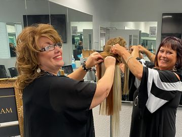 Two women styling a blonde hairpiece in a salon.