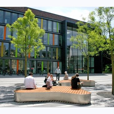 People sitting and walking near modern benches and green trees outside a colorful building. bancas de acero inoxidable
papeleras
paradas