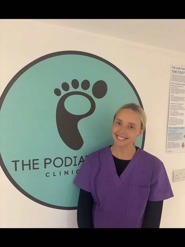 Smiling podiatrist in purple scrubs stands by clinic logo on wall.