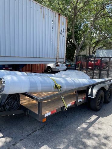 A large rolled item secured on a trailer with a yellow strap near a shipping container.
