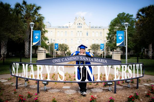 Graduate posing with St. Mary's University sign and campus building.