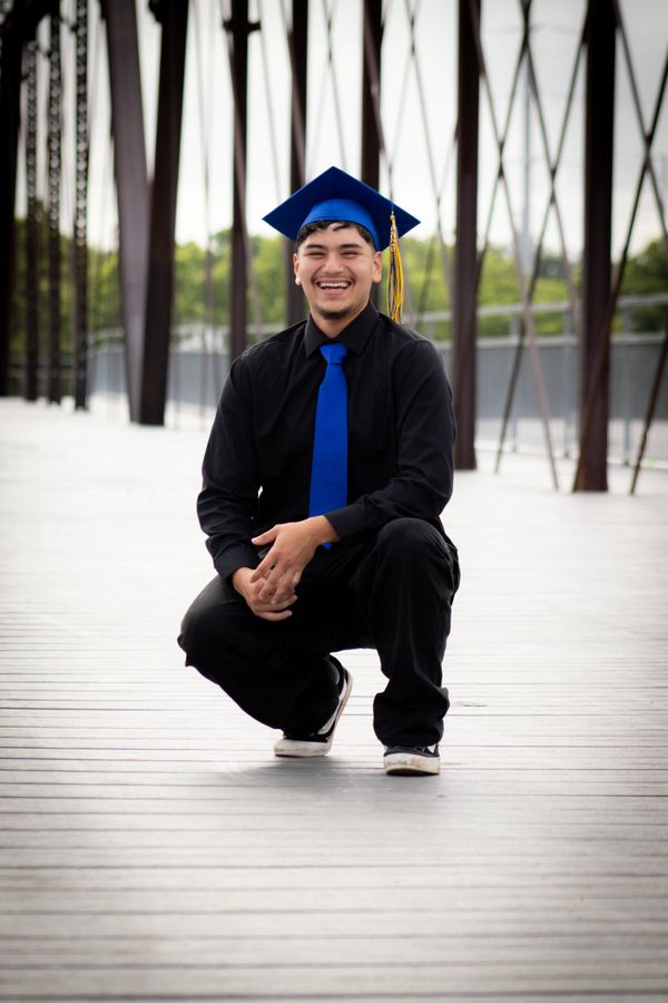 Young graduate kneeling on a bridge, smiling in cap and gown.