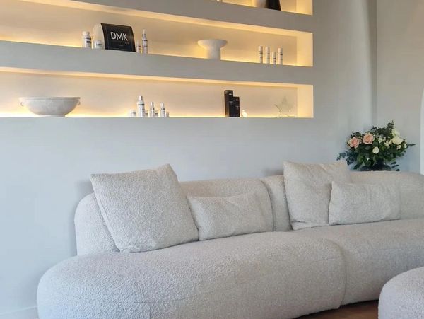 Modern beige sofa with cozy cushions beneath softly lit wall shelves displaying skincare products.