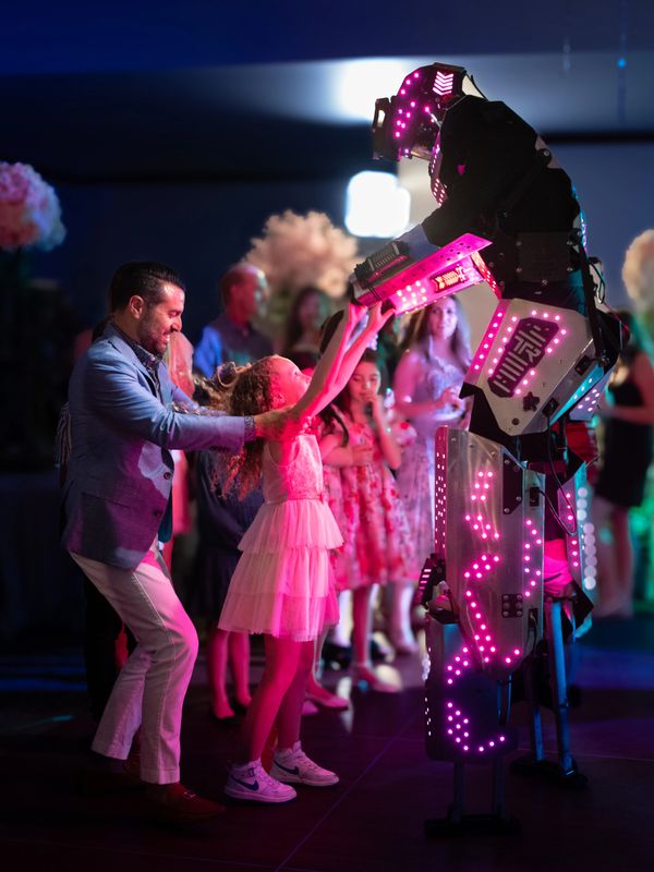 A man and a girl interact joyfully with a glowing LED robot at a party.