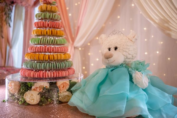 A teddy bear in a blue dress next to a colorful macaron tower with flowers underneath.