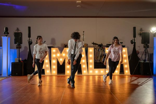 Three young people dancing in front of illuminated letters spelling AMALIA.