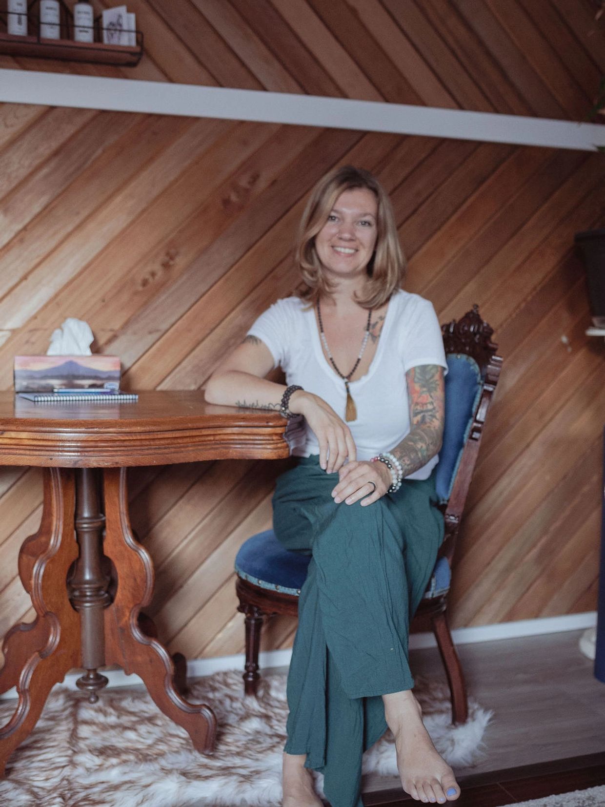 Smiling woman with tattoos sitting barefoot on an ornate chair beside a wooden table.