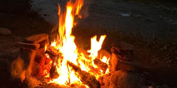 A campfire burning brightly by a river at dusk in a forested area.