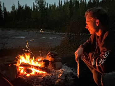 A man gazes thoughtfully into a campfire at dusk by a forest river.