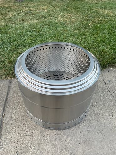 A stainless steel fire pit with perforated inner walls on a concrete surface.