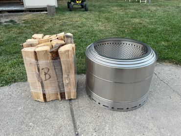 Stacked firewood and a shiny metal fire pit on a concrete surface near grass.