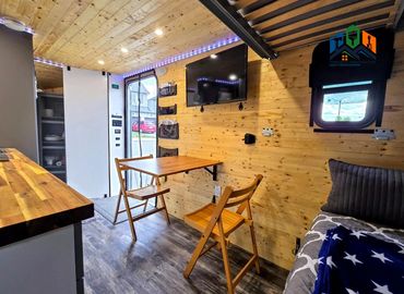 Pine Box camper corner view showing dining area with table, door, window, TV & natural light