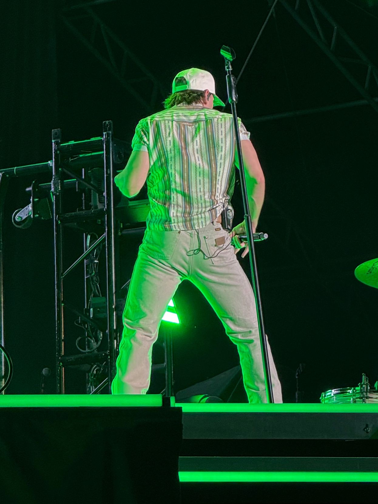 Performer in white pants and cap on stage with drummer under green lights.