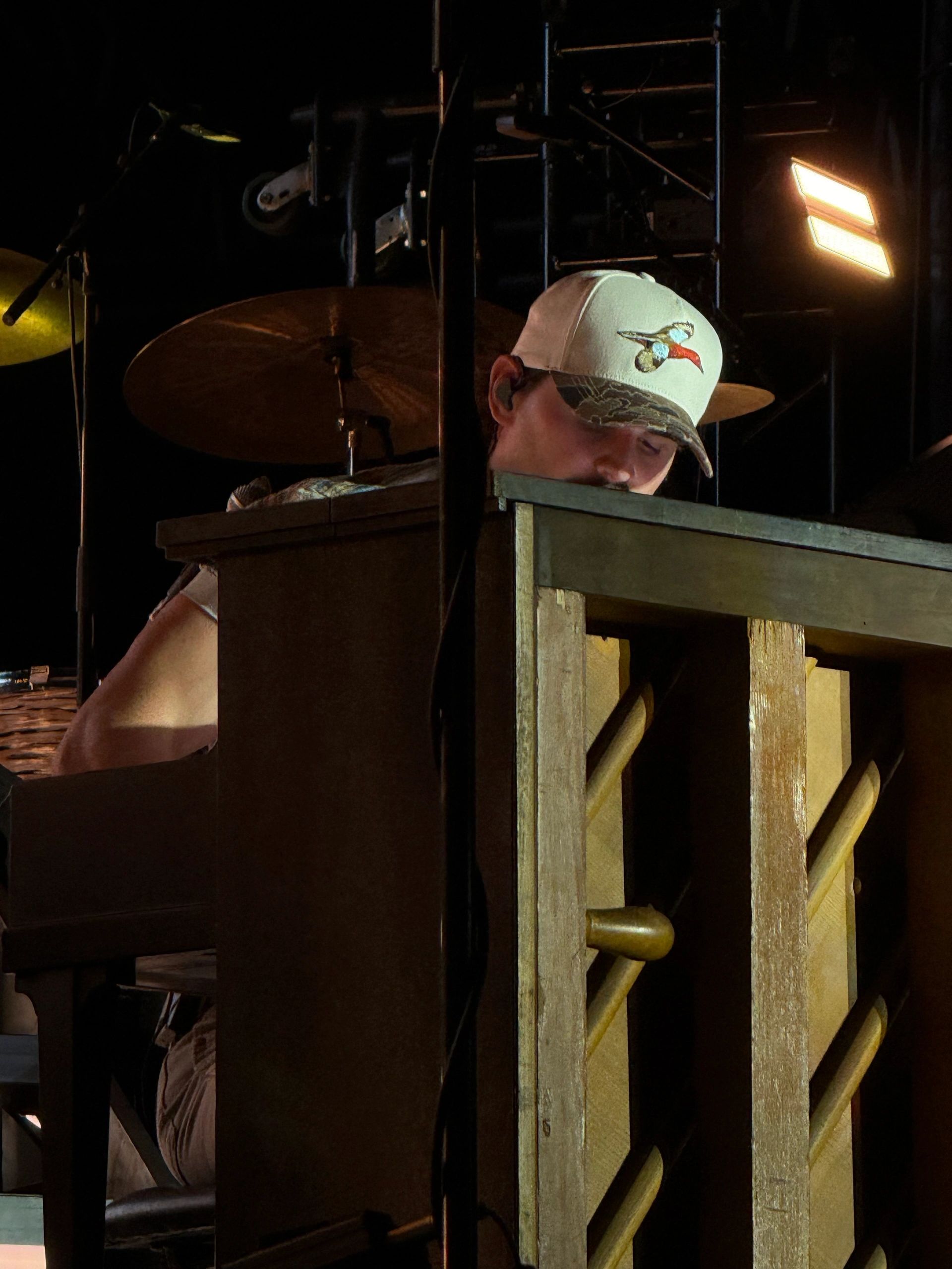 Musician in a camo hat plays piano on dimly lit stage.