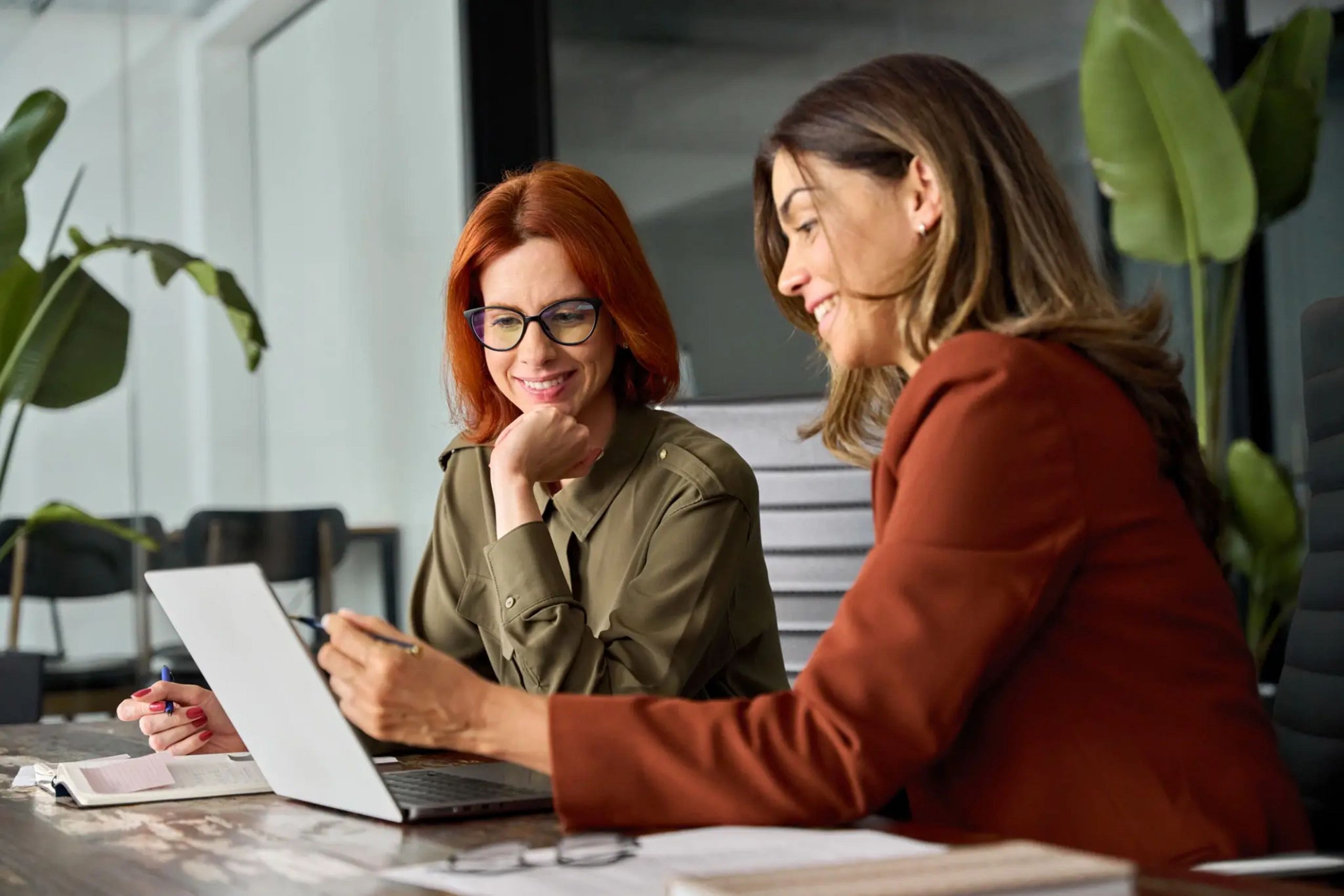 Two women smiling and working together on a laptop in a modern office.