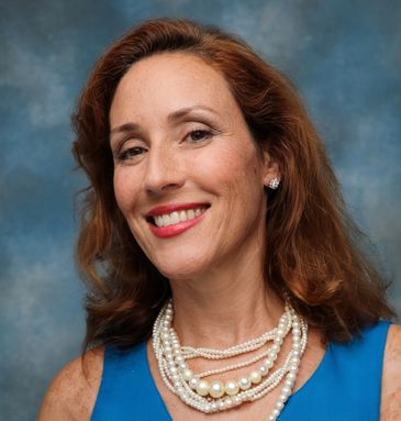 Smiling woman with red hair wearing a blue dress and pearl necklaces.
