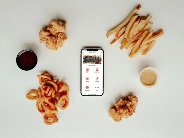 Smartphone surrounded by fried chicken pieces, curly fries, regular fries, and dipping sauces on a white surface.
