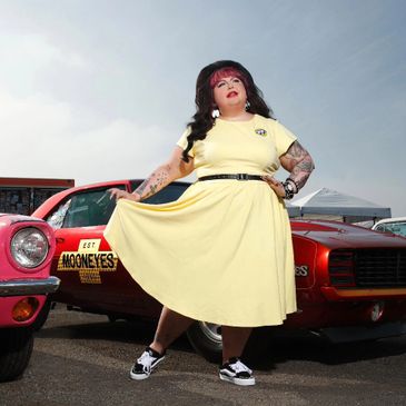 Person in yellow dress posing between two classic red cars.