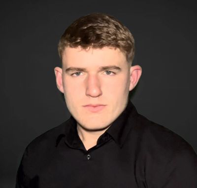 Rio Kavanagh with short hair wearing a black shirt against a dark background.