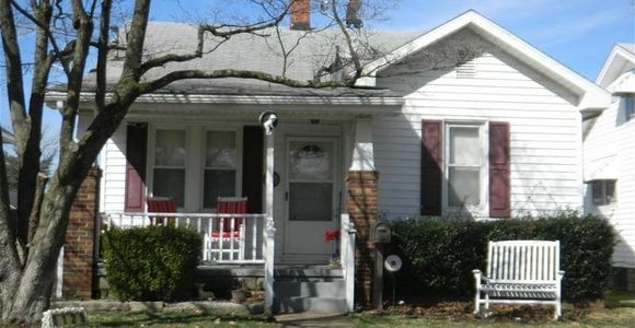 A small white house with a porch and outdoor seating under a tree.