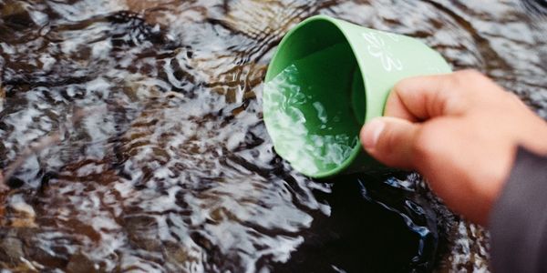 Hand holding green cup scooping water from a flowing stream.