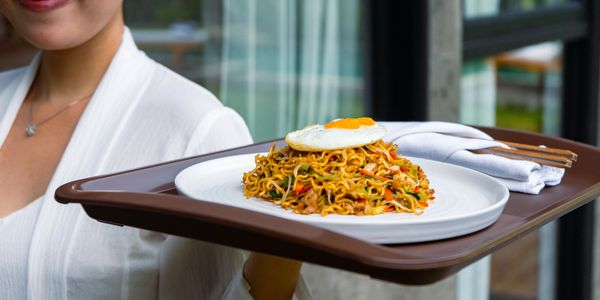 A person holding a tray with a plate of noodles topped with a fried egg.