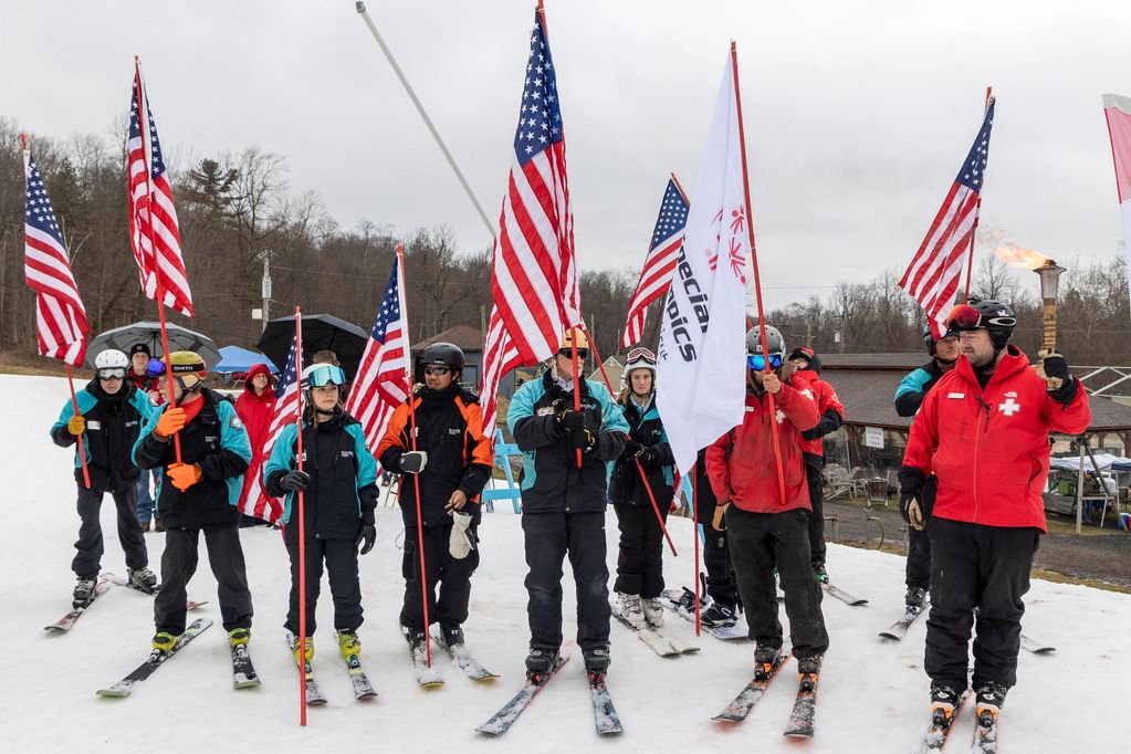 Skiers holding American flags and a torch on a snowy slope during a ceremony.