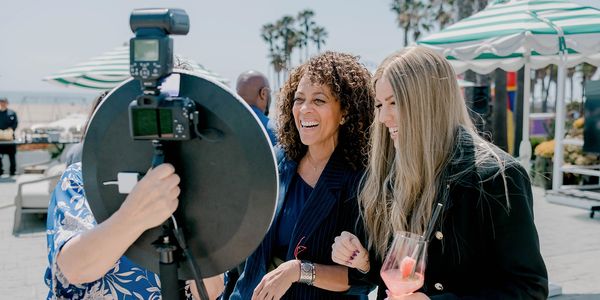 Two women smiling and posing for a photo outdoors with a camera and reflector.
