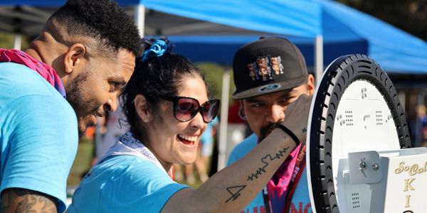 Three people smile and engage with a spinning prize wheel at an outdoor event.