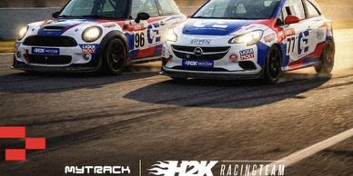 Two race cars with H2K Racing Team livery on a track at sunset.