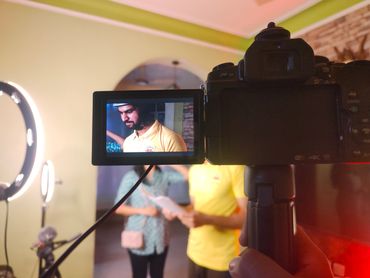 Camera screen showing a man in a yellow shirt and helmet being filmed indoors.