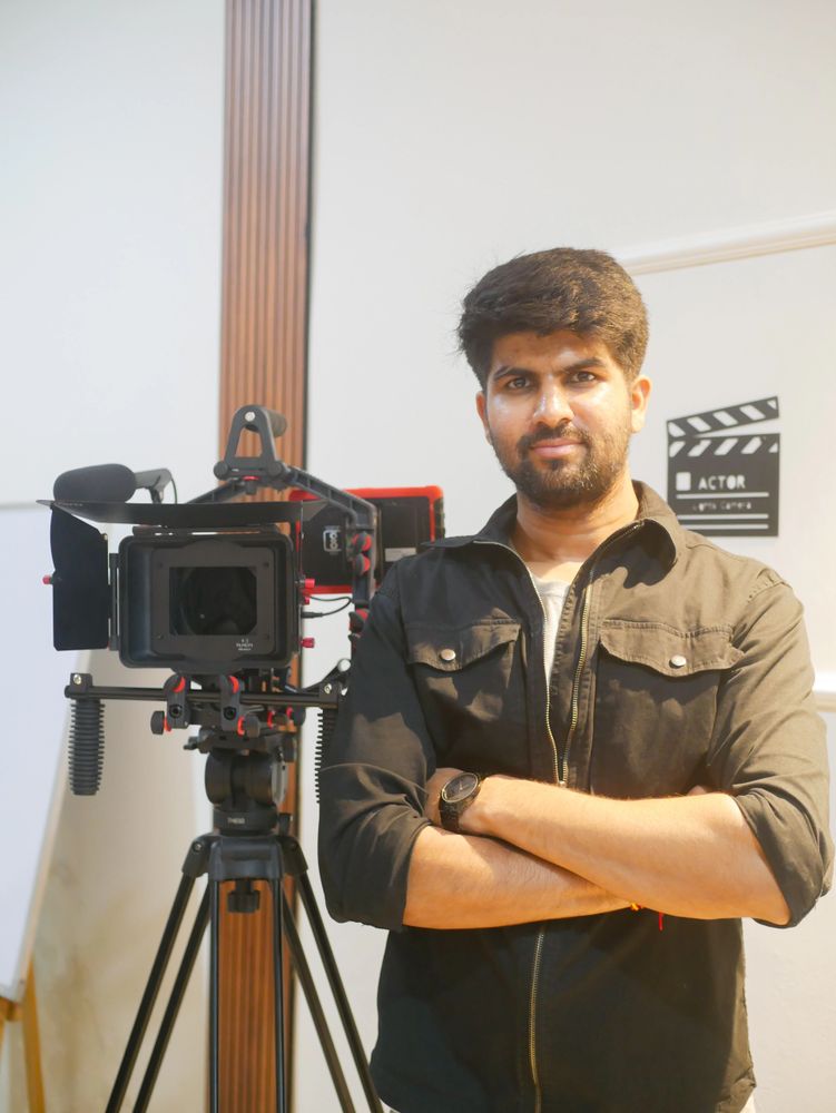 Man standing confidently beside professional video camera on tripod indoors.