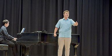 A young man sings on stage with piano accompaniment.