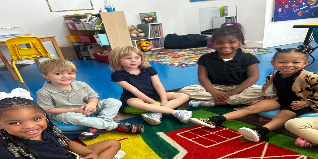 Five children sitting in a circle on a colorful classroom rug, smiling.