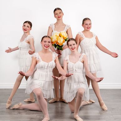 Five ballerinas posing in white dresses with one holding flowers.