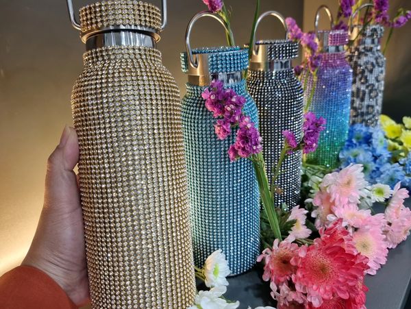 Hand holding a gold rhinestone water bottle with decorated bottles and flowers in the background.