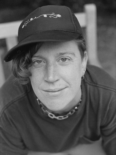Black-and-white portrait of a person wearing a cap and chain necklace, smiling gently.