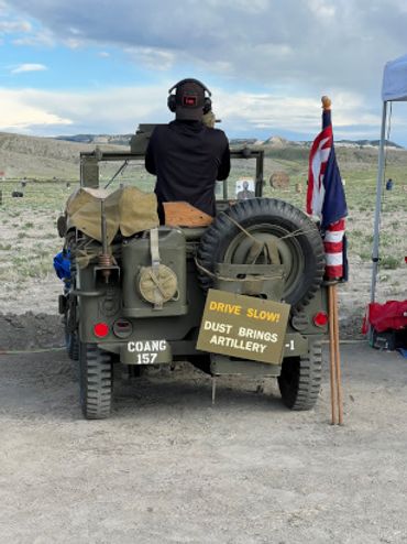 Person aiming rifle from a vintage military jeep in a desert setting.