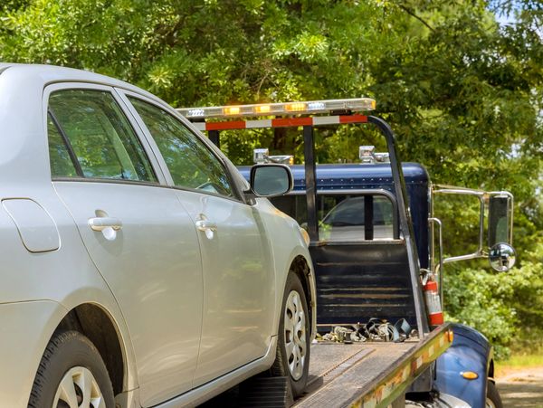 A silver car is being loaded onto a tow truck in a sunny, green outdoor