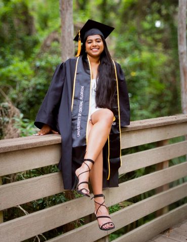 Smiling graduate in cap and gown sitting on a wooden fence.