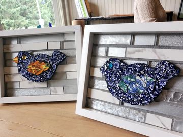 two framed mosaics one with a blue bird and the other with a blue and orange bird against a tile bac