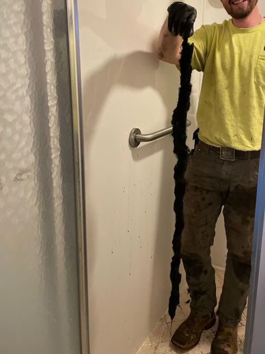 Man holding a long, dirty black object inside a bathroom shower.