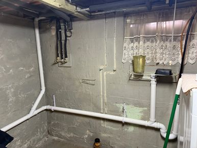 Basement corner with exposed pipes and a small window with lace curtains.
