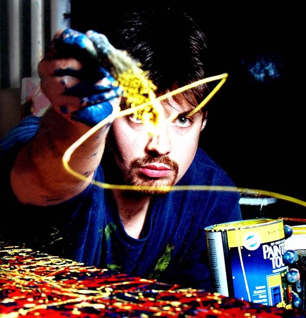 The artist throws paint on canvas while staring into the camera