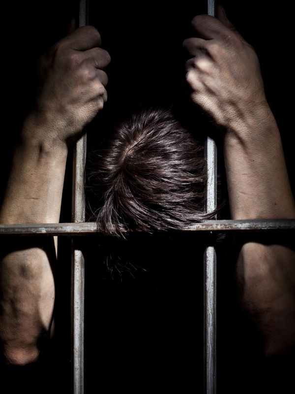 Person gripping prison bars with head down in darkness.