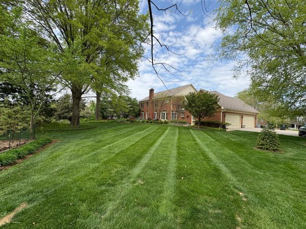 A large brick house with a manicured lawn and tall trees under a partly cloudy sky.