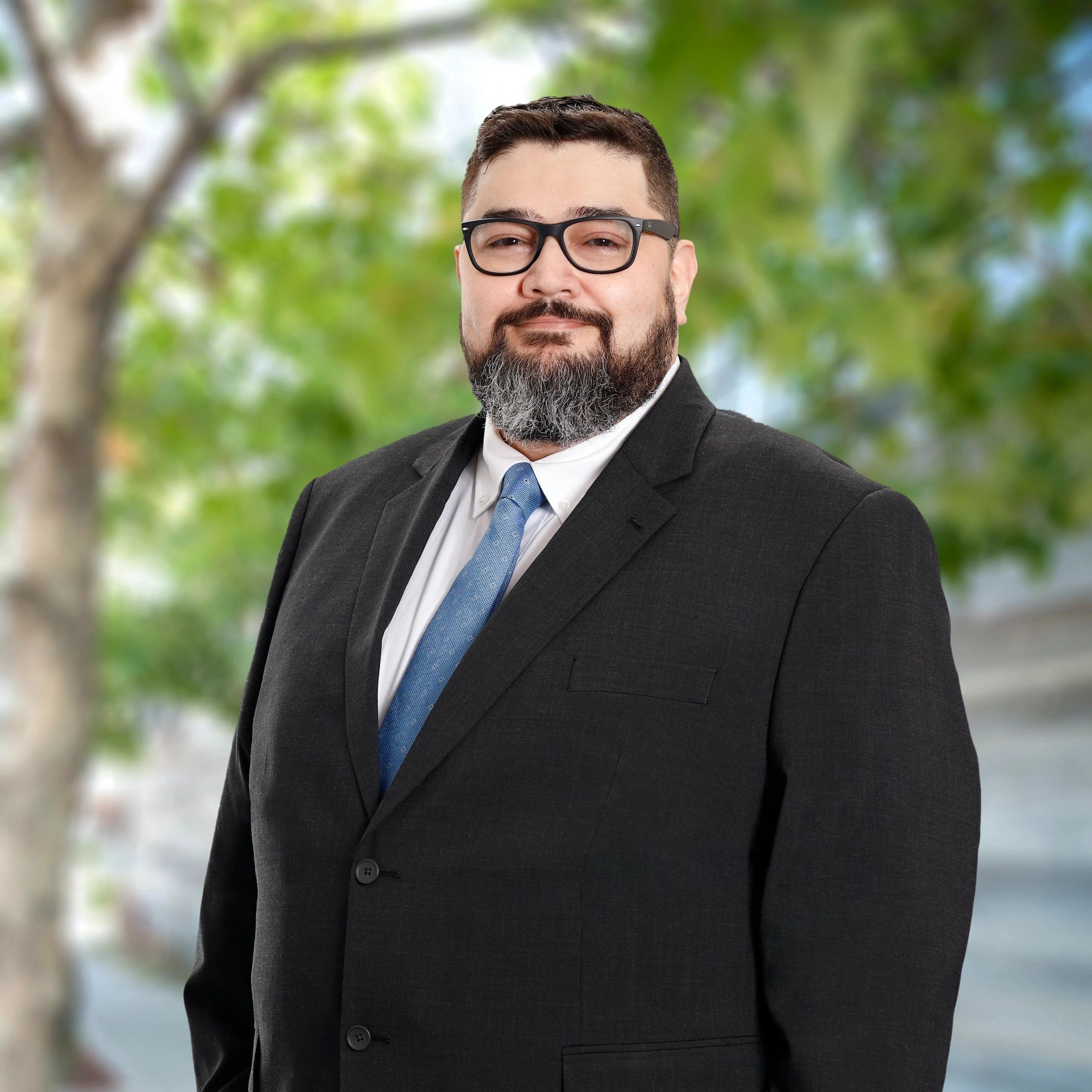 Man in suit with glasses and a beard stands outdoors.