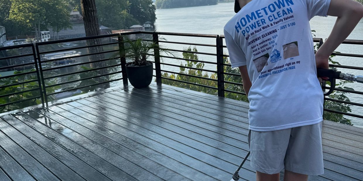 Person power washing a wooden deck overlooking a serene lake.