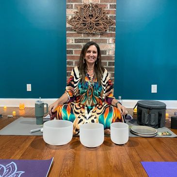 Woman in colorful dress sitting with crystal singing bowls in a meditation room.