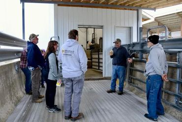 A group of people listening to a man speaking inside a barn-like structure.